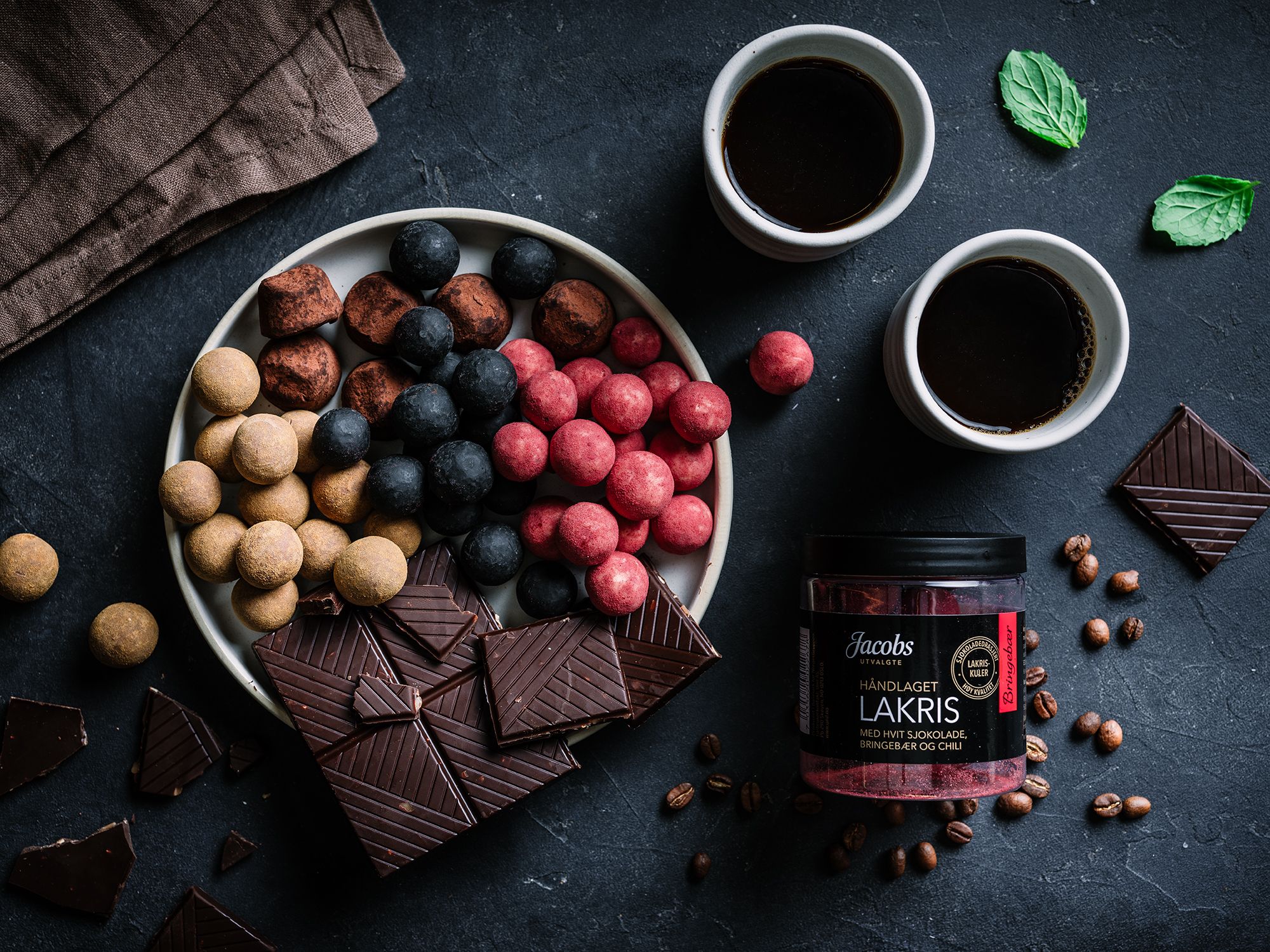 En skål med ulike typer sjokoladebiter og biter av mørk sjokolade, sammen med to kopper kaffe og en liten krukke med Jacobs Lakris, på en mørk overflate.