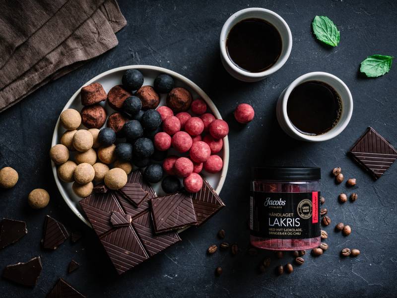 En skål med ulike typer sjokoladebiter og biter av mørk sjokolade, sammen med to kopper kaffe og en liten krukke med Jacobs Lakris, på en mørk overflate.
