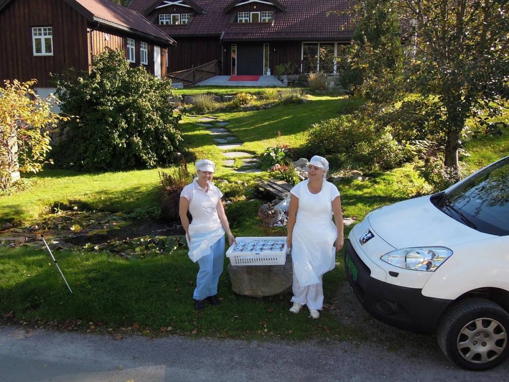 To personer i hvite klær og hårnetter står ved et bord utendørs i en hage, med en hvit bil parkert ved siden av og en stor hus i bakgrunnen.