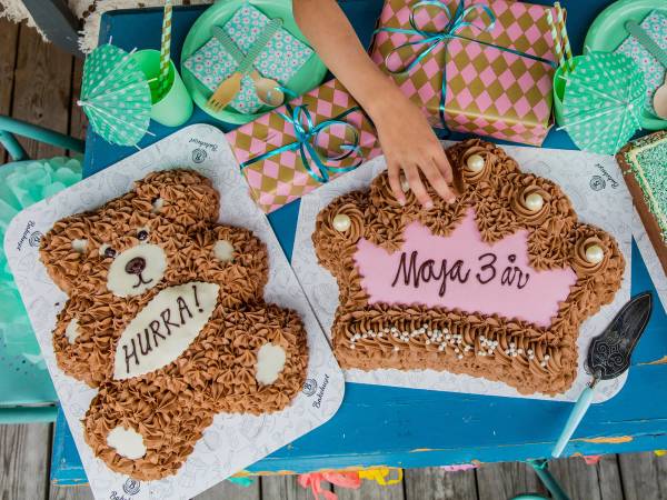 To kaker, en med en bjørnefigur og en med teksten 'Maja 3 år', omgitt av gaveesker og dekorasjoner, på et bord utendørs.