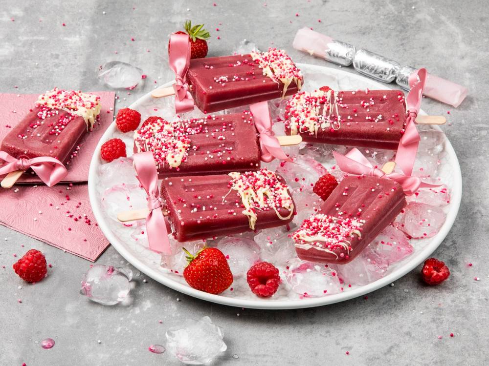 Små rosa og hvite sjokoladebarer pyntet med strøssel på en hvit tallerken, omgitt av friske jordbær og små dekorative elementer.