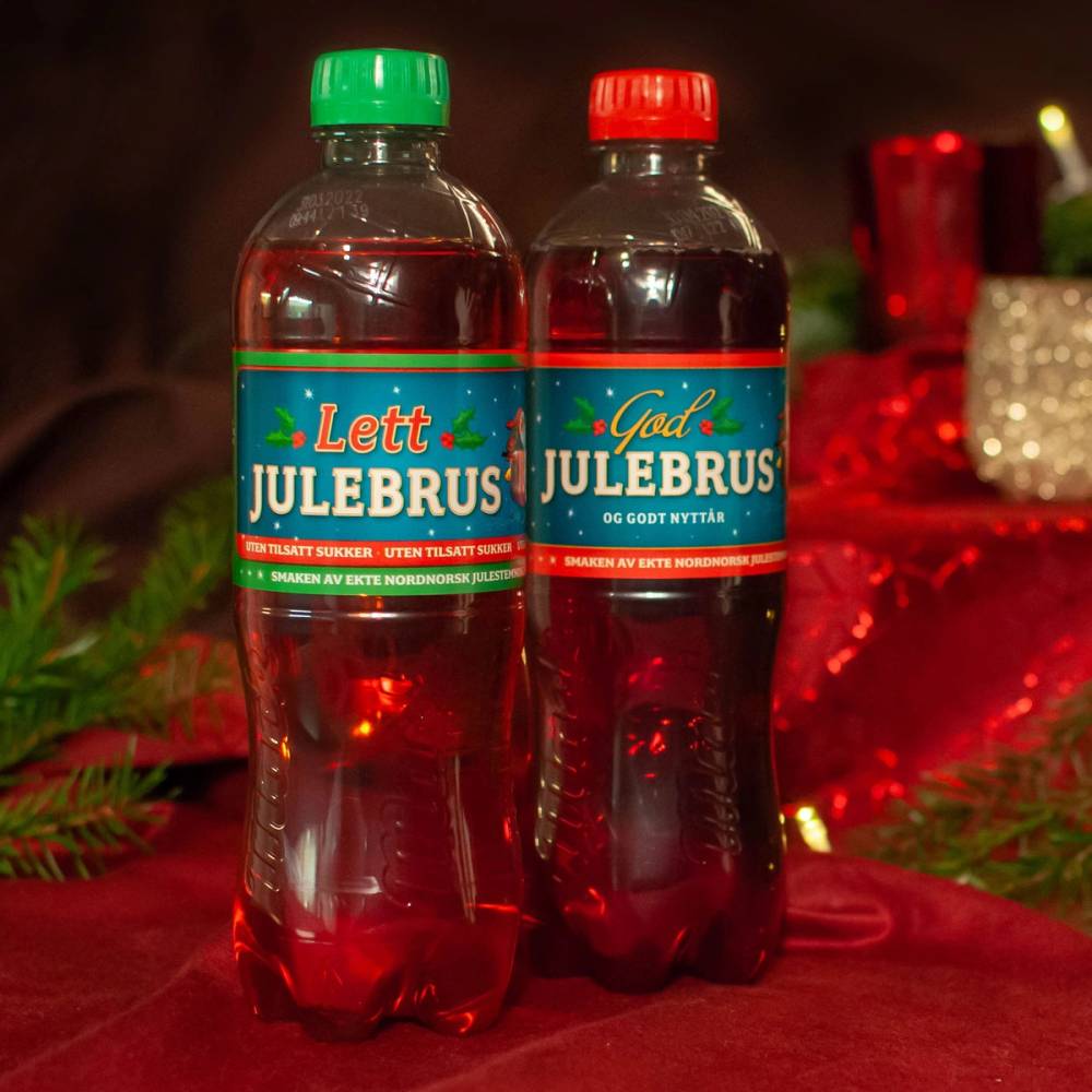 To plastflasker med julebrus, en med grønn kork og en med rød kork, står på et bord med julepynt rundt.