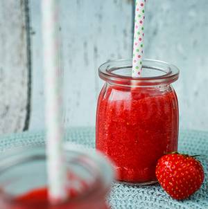 En glass med jordbærslush med et sugerør, og en hel jordbær ved siden av på et bord.