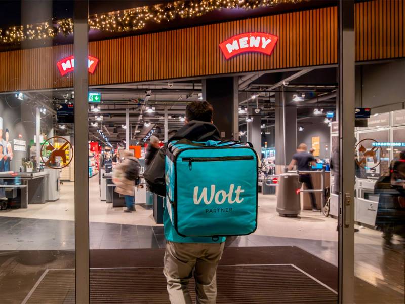 En person med en Wolt-matleveringsveske går inn i en butikk eller restaurant, sett fra bakfra.
