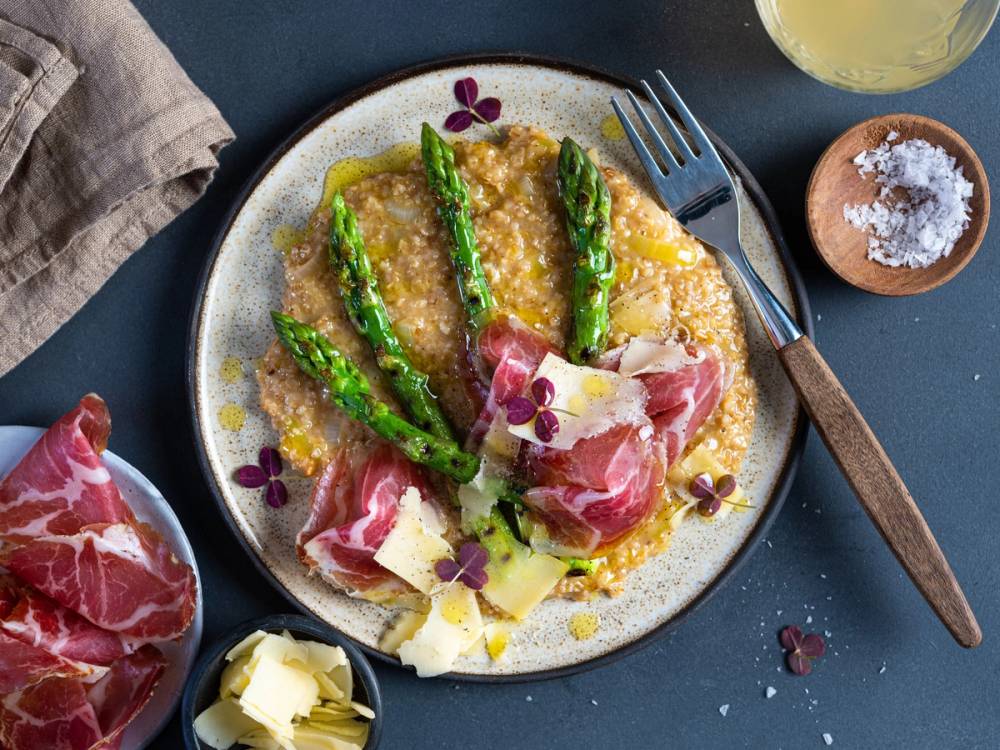 Asparges med skinke og ost på en tallerken, omgitt av en liten bolle med salt og et glass med en lys drikke