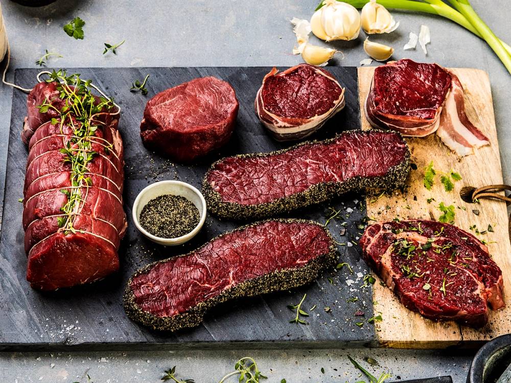 Ulike biter av grillet biff og kjøtt, med en liten skål med krydder og noen hvitløksfedd i bakgrunnen, på en stekeplate.