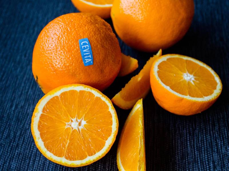 Ferske appelsiner, hvorav noen er hel og andre er skåret opp, med en Cevita-merket appelsin på en mørk bakgrunn.