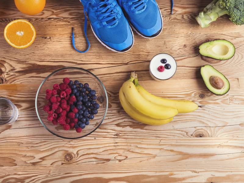 Frukt og treningsutstyr på et trebord, inkludert bananer, avokado, bær og blå joggesko.