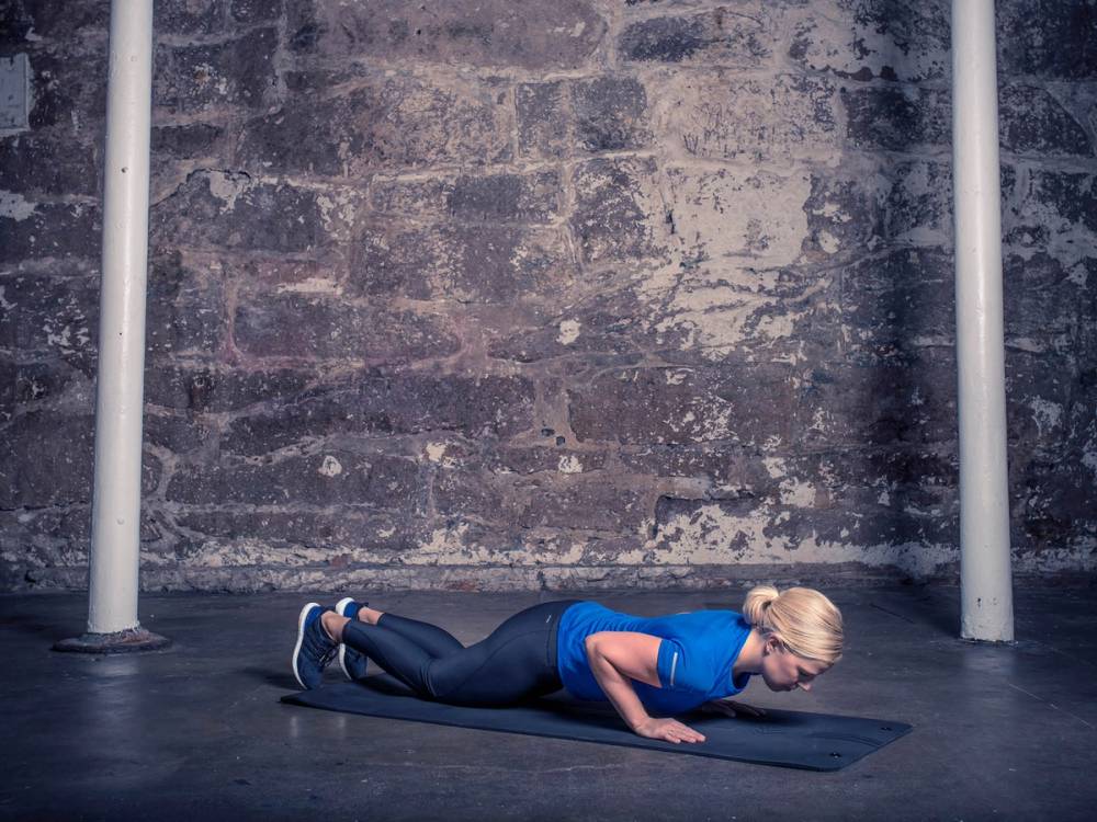 En kvinne utfører smale pushups på gulvet i et treningsrom med en murvegg i bakgrunnen.