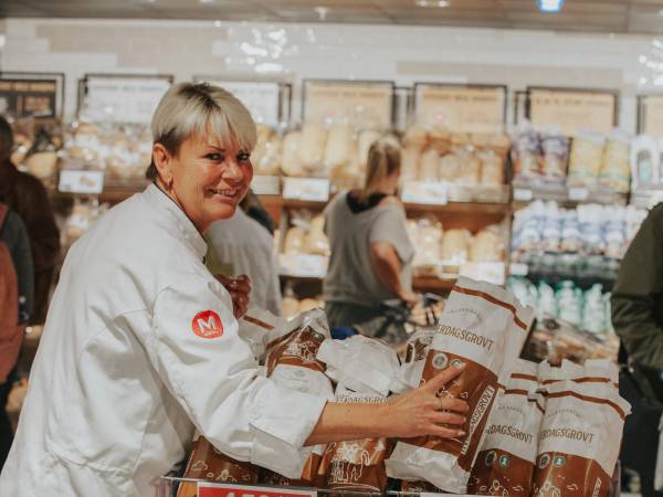 En person med kledd i hvit frakk handler i en dagligvarebutikk med hyller fylt med matvarer i bakgrunnen.