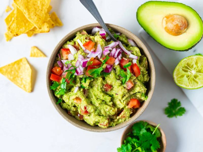 En bolle med guacamole omgitt av tortillachips, avokado, lime og frisk koriander på et hvitt bord
