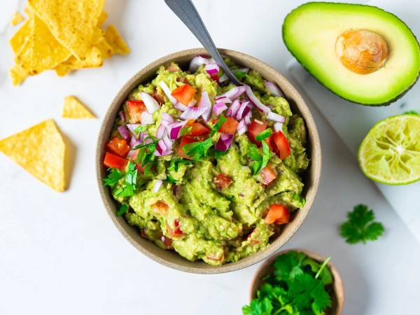 En bolle med guacamole omgitt av tortillachips, avokado, lime og frisk koriander på et hvitt bord