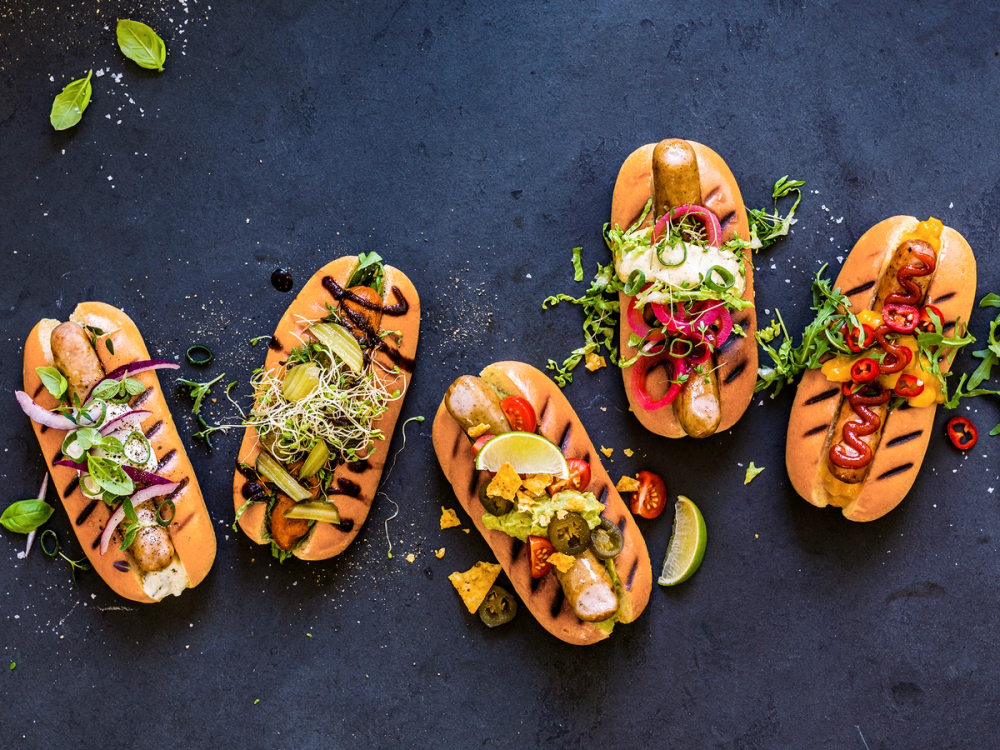 Fem pølser med ulike toppings og garnityr på en mørk bakgrunn, klare for grilling eller servering.