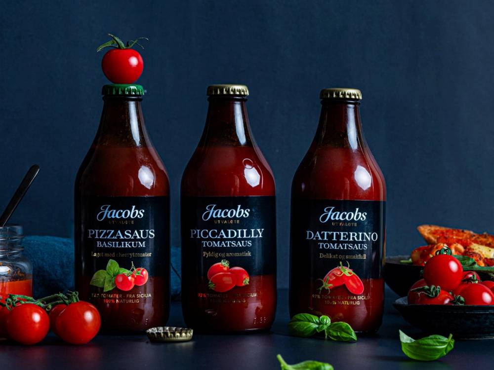 Tre flasker med tomatsaus fra Jacobs, omgitt av friske tomater og basilikumblad på en mørk bakgrunn.