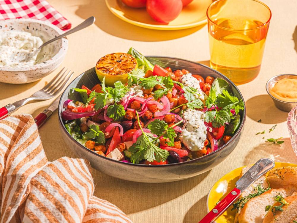 En fargerik salat med grønnsaker, urter og ost i en svart bolle på et bord med ulike retter og drikkevarer rundt.