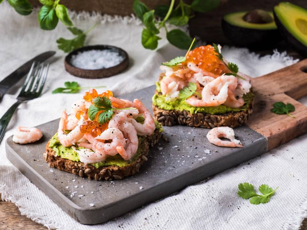 Rekesmorsbrød med avokado og reker på en steinplate, pyntet med urter og sitron, omgitt av avokado og grønne blader.