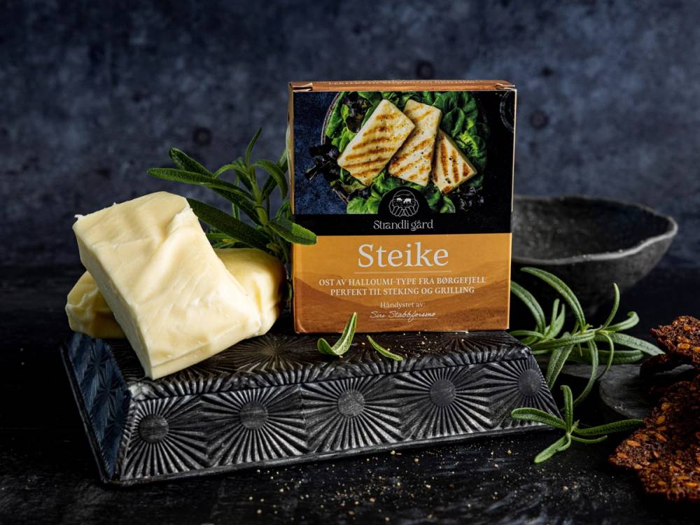 Steike ost fra Strandli Gård med urter og brød i bakgrunnen, presentert på en svart mønstret tallerken.