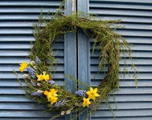 En påskekrans laget av grener med gule påskeliljer og lilla blomster, hengt opp på en blå vegg med loddrette paneler.