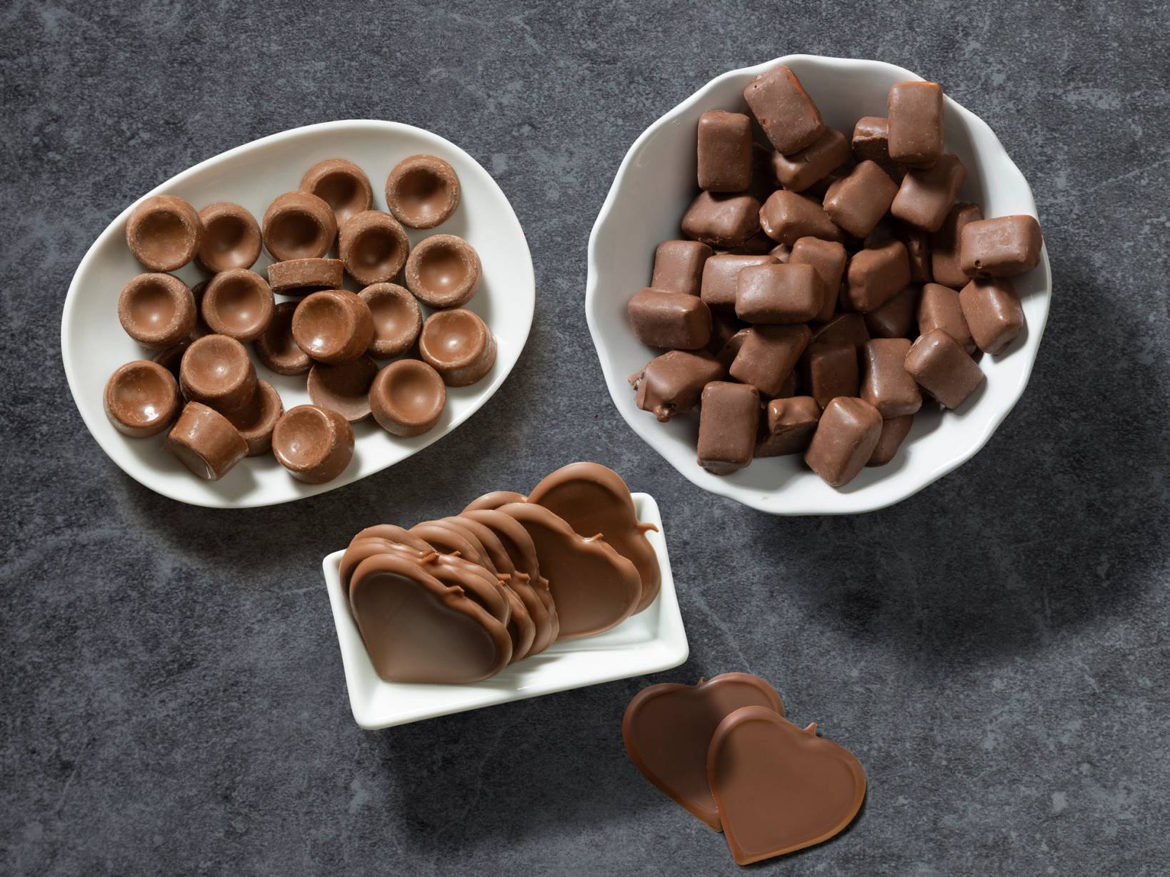 Ulike typer sjokolade i små skåler på en mørk overflate, inkludert sjokoladeknapper, terninger og sjokoladekjeks.