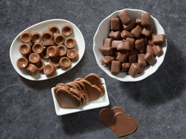 Ulike typer sjokolade i små skåler på en mørk overflate, inkludert sjokoladeknapper, terninger og sjokoladekjeks.