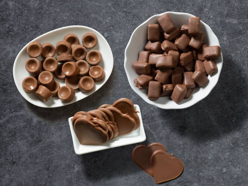 Ulike typer sjokolade i små skåler på en mørk overflate, inkludert sjokoladeknapper, terninger og sjokoladekjeks.