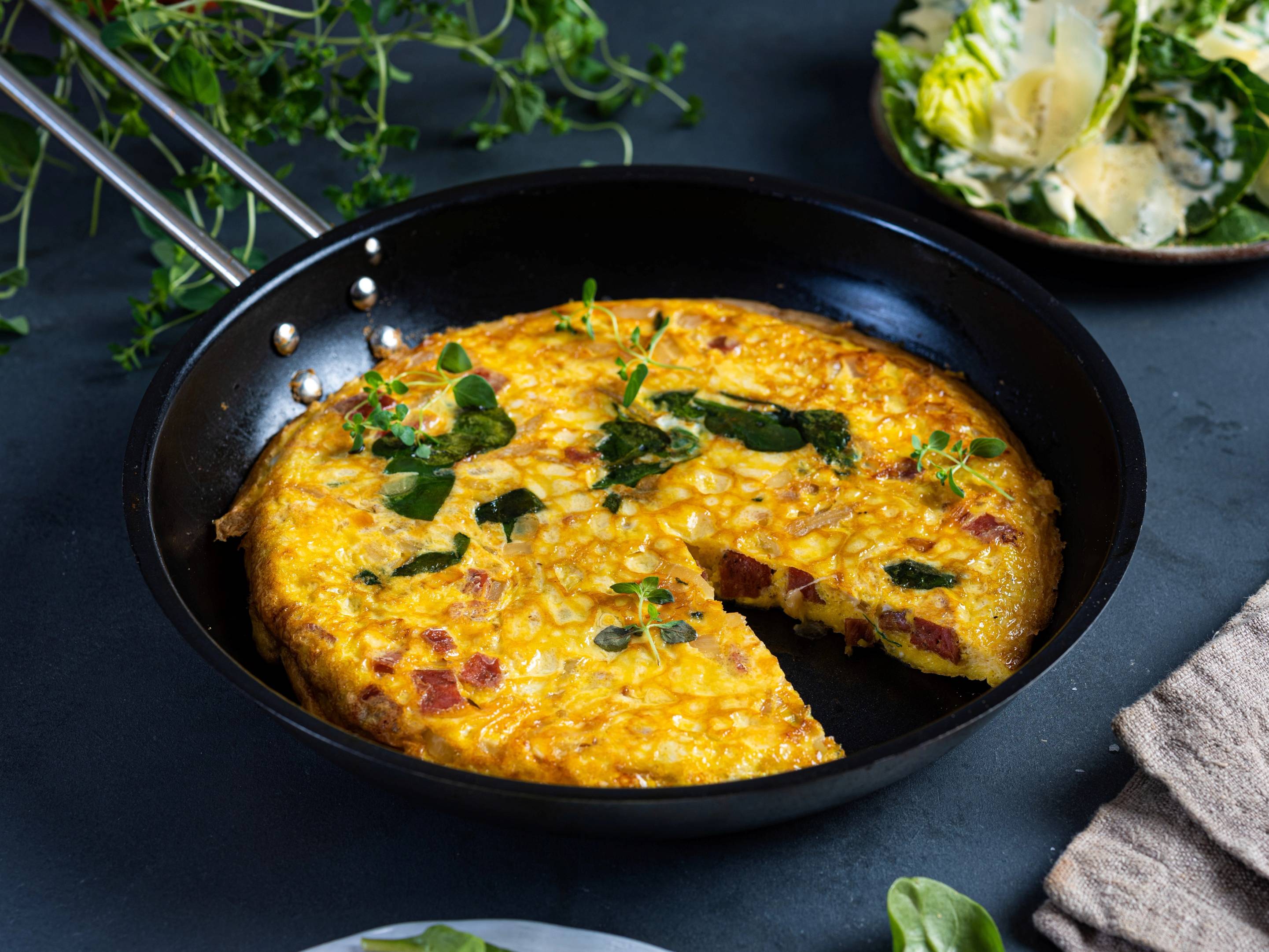Bondeomelett med grønnsaker og urter i en stekepanne, med en tallerken med blomster i bakgrunnen.
