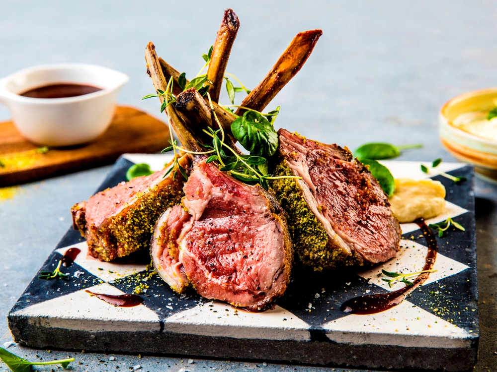 Lammecarré med urteskorpe servert med grønnsaker og saus på en tallerken.