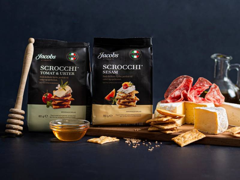 To pakker med Jacobs utvalgte Scrocchi kjeks, en skje med honning, og ulike oster og kjøtt på et skjærebrett, mot en mørk bakgrunn.