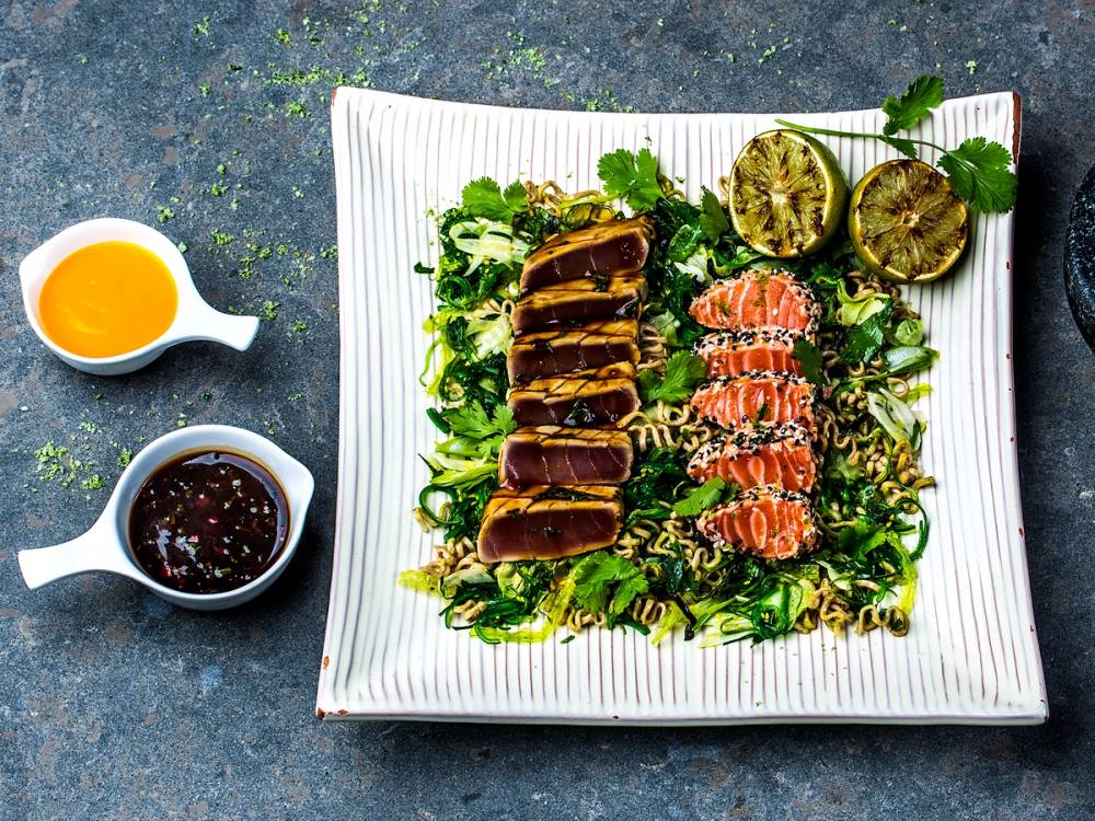 Sushibiter med tunfisk tataki, servert med lime, sitron og soyasaus på en hvit firkantet tallerken, med en liten skål med soyasaus og en skål med en gul saus ved siden av.