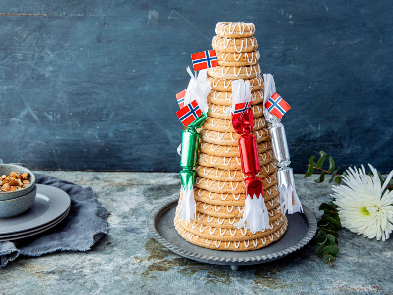 En kransekake pyntet med små norske flagg og røde og grønne dekorasjoner på en sølvfat, plassert på et bord med en blå bakgrunn.