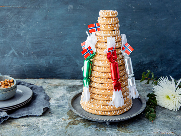 En kransekake pyntet med små norske flagg og røde og grønne dekorasjoner på en sølvfat, plassert på et bord med en blå bakgrunn.