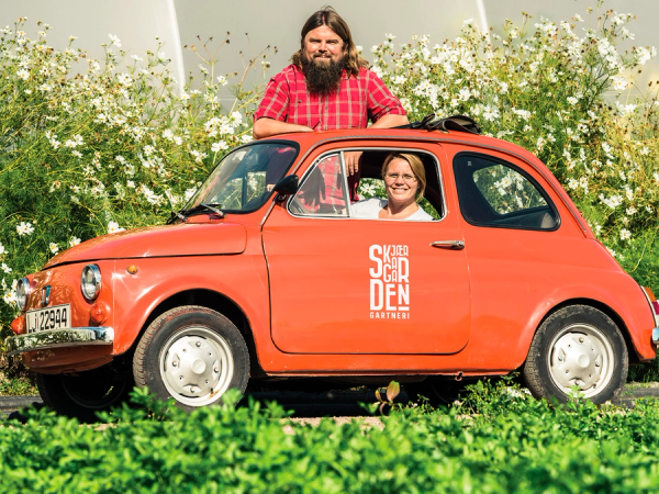 To personer i en liten rød bil med teksten 'SØNDEN' på siden, parkert foran en blomstereng med hvite blomster, en av personene sitter i førersetet og den andre står ved siden av bilen.