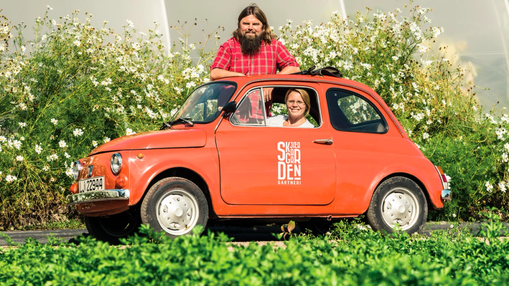 To personer i en liten rød bil med teksten 'SØNDEN' på siden, parkert foran en blomstereng med hvite blomster, en av personene sitter i førersetet og den andre står ved siden av bilen.