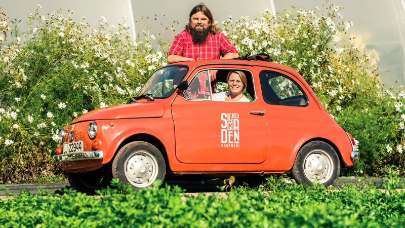 To personer i en liten rød bil med teksten 'SØNDEN' på siden, parkert foran en blomstereng med hvite blomster, en av personene sitter i førersetet og den andre står ved siden av bilen.