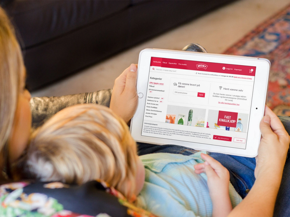 En person holder en nettbrett som viser en nettside med en meny og ulike tilbud, med en liten barn som ser på sammen.