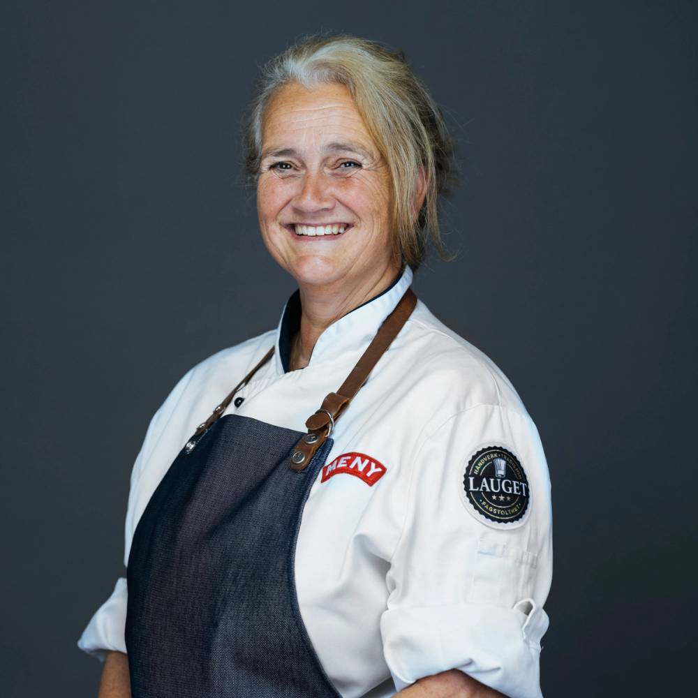 En kvinne i kokkeuniform med en logo på armen og en hvit jakke, står mot en mørk bakgrunn.