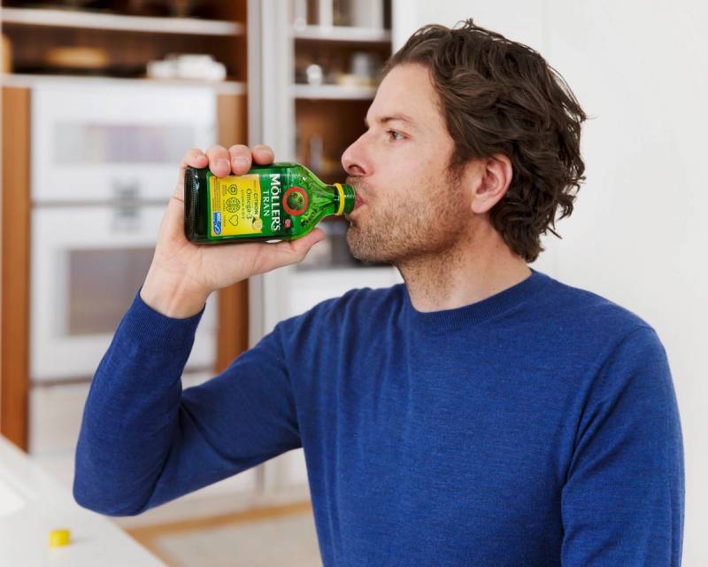 Person som drikker tran fra en flaske i et kjøkken, med kjøkkeninnredning i bakgrunnen.