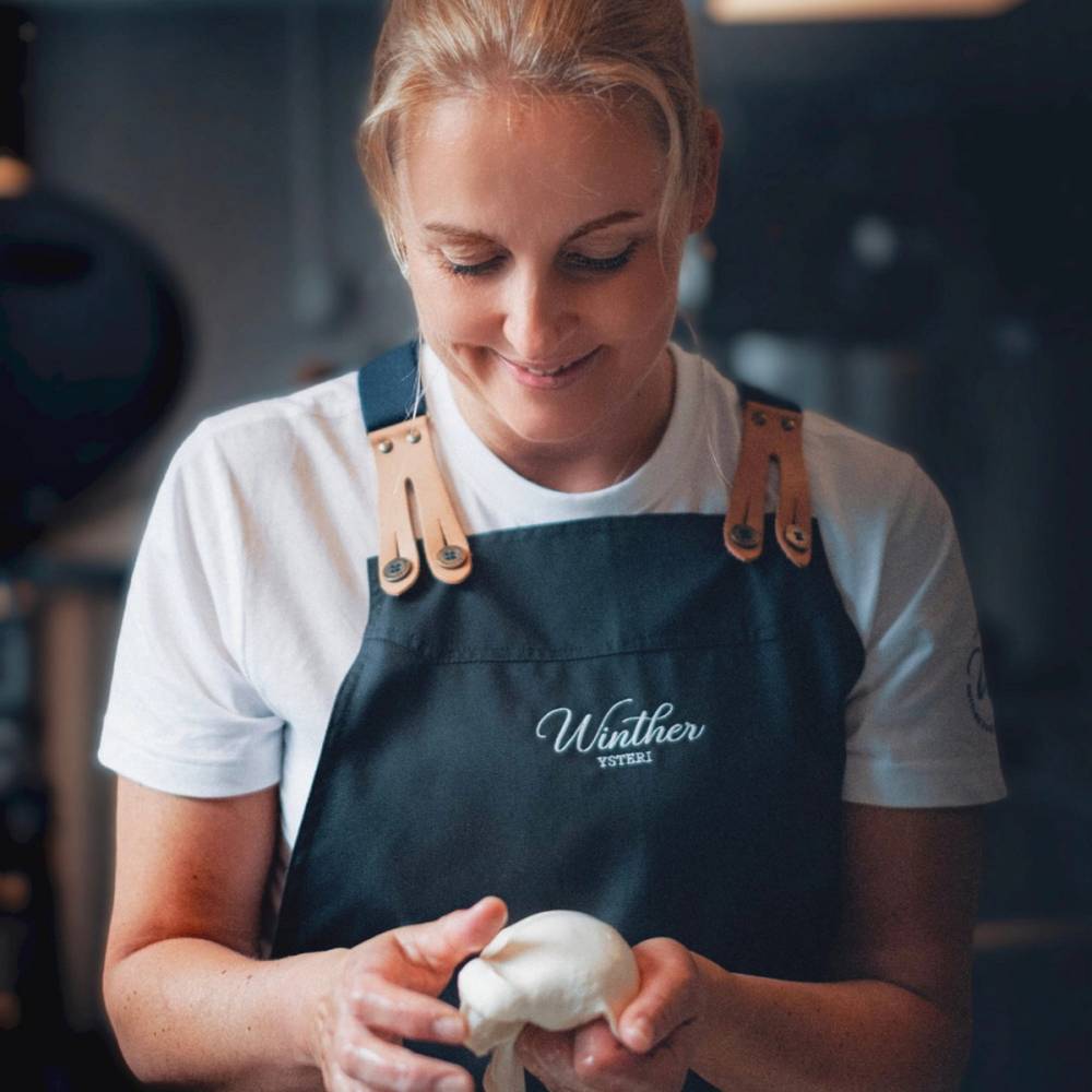 En person iført hvit t-skjorte og blå forkle som holder en ball av deig i hendene, i et kjøkkenmiljø.