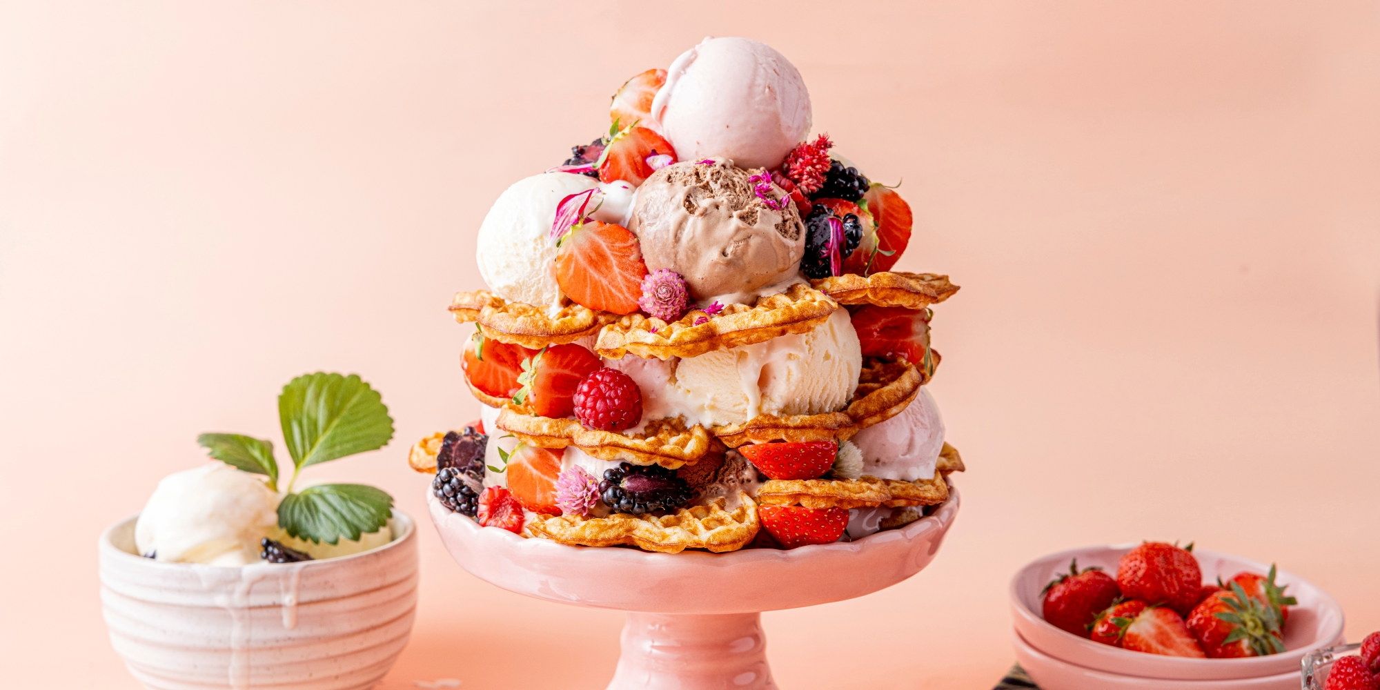 Et vaffeltårn med iskrem og friske bær på en rosa tallerken, omgitt av små skåler med mer bær og mynteblader.