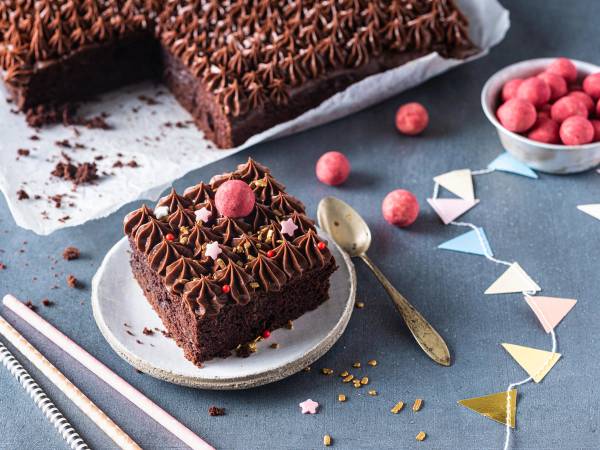 En langpanne med lys sjokoladekrem og pyntet med sjokoladekremstjerner og en rosa marsipankule, med et stykke av kaken på en liten tallerken og en skje ved siden av.