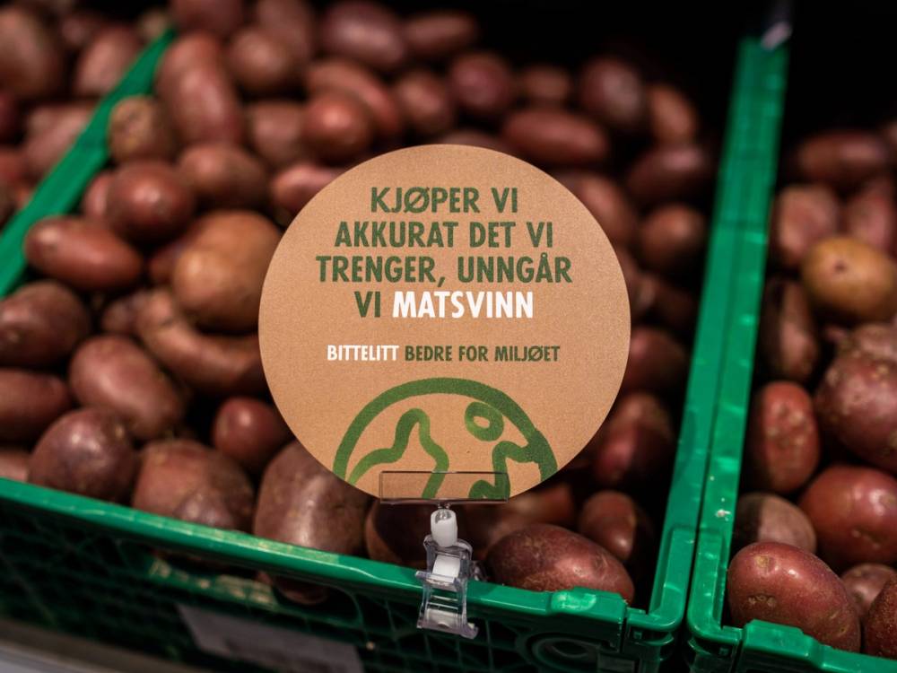 En plastkasse med poteter og et rundt skilt med teksten 'KJØPER VI AKKURAT DET VI TRENGER, UNNGÅR VI MATSVINN' og en grønn illustrasjon av en hånd og en tallerken.