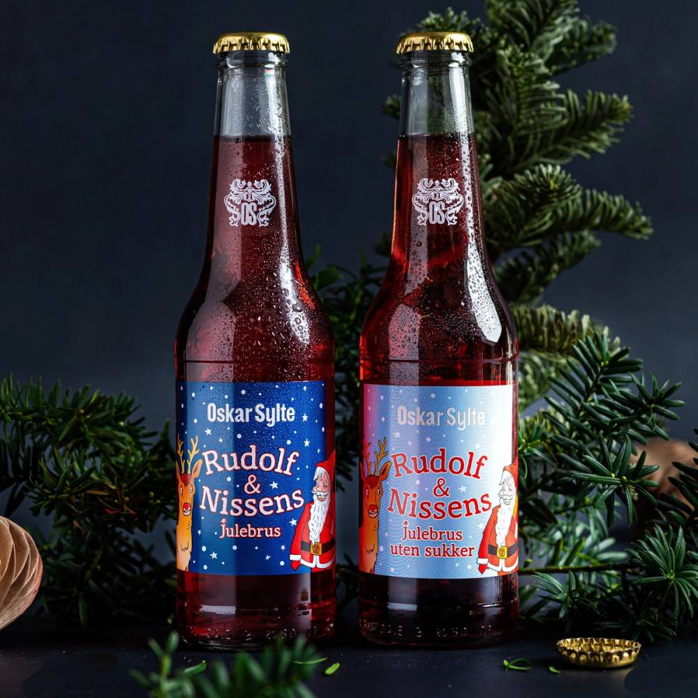 To flasker julebrus med etiketter for Rudolf og Nissens julebrus, plassert foran julegrener og pynt.