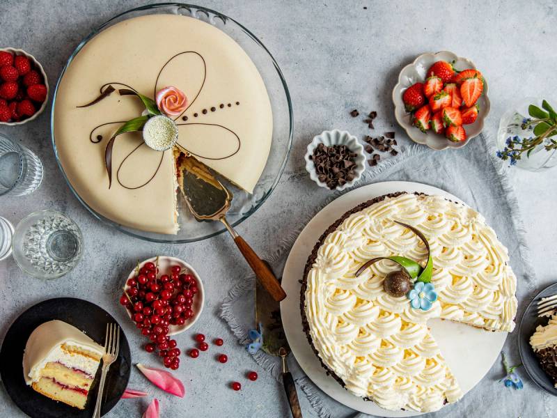 To kaker, en med hvit glasur og dekorasjon, og en med krem og frukt, på et bord med bær og blomster rundt.