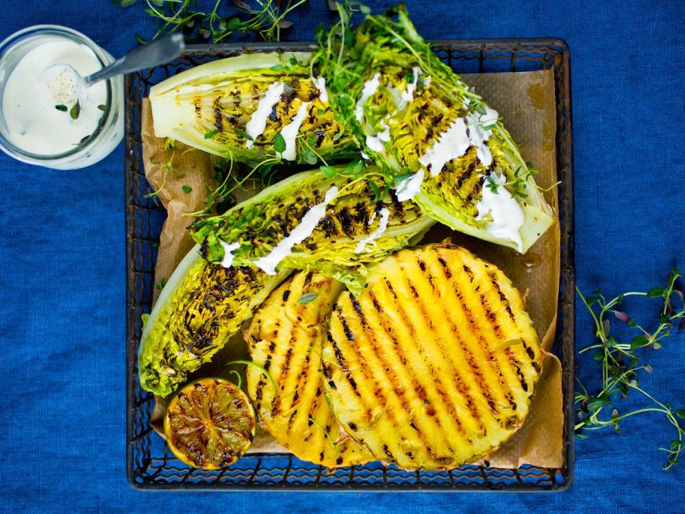 Grillet hjertesalat med mormordressing og grillede maiskolber på en serveringsfat med en sitronhalvdel og urter rundt.