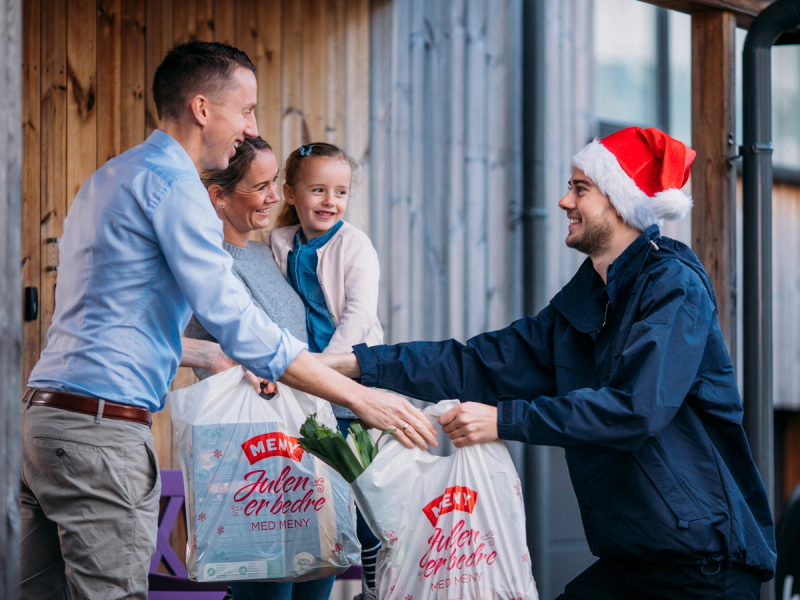 En person i julehatt gir en gave til en annen person i en butikk eller på et arrangement, med to andre personer i bakgrunnen.