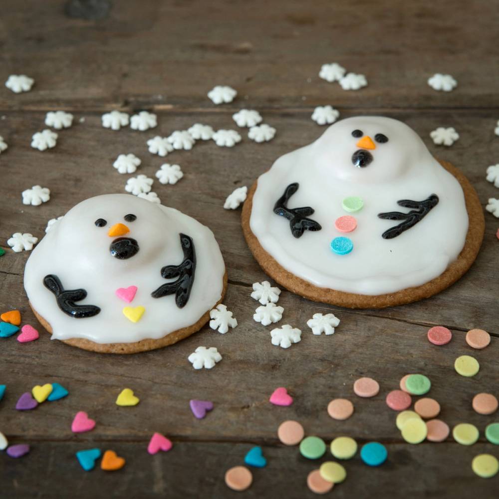 To pepperkaker pyntet som snømenn med hvit glasur, svarte armer, øyne og munn, omgitt av små fargede konfetti og snøfnugg av sukker.