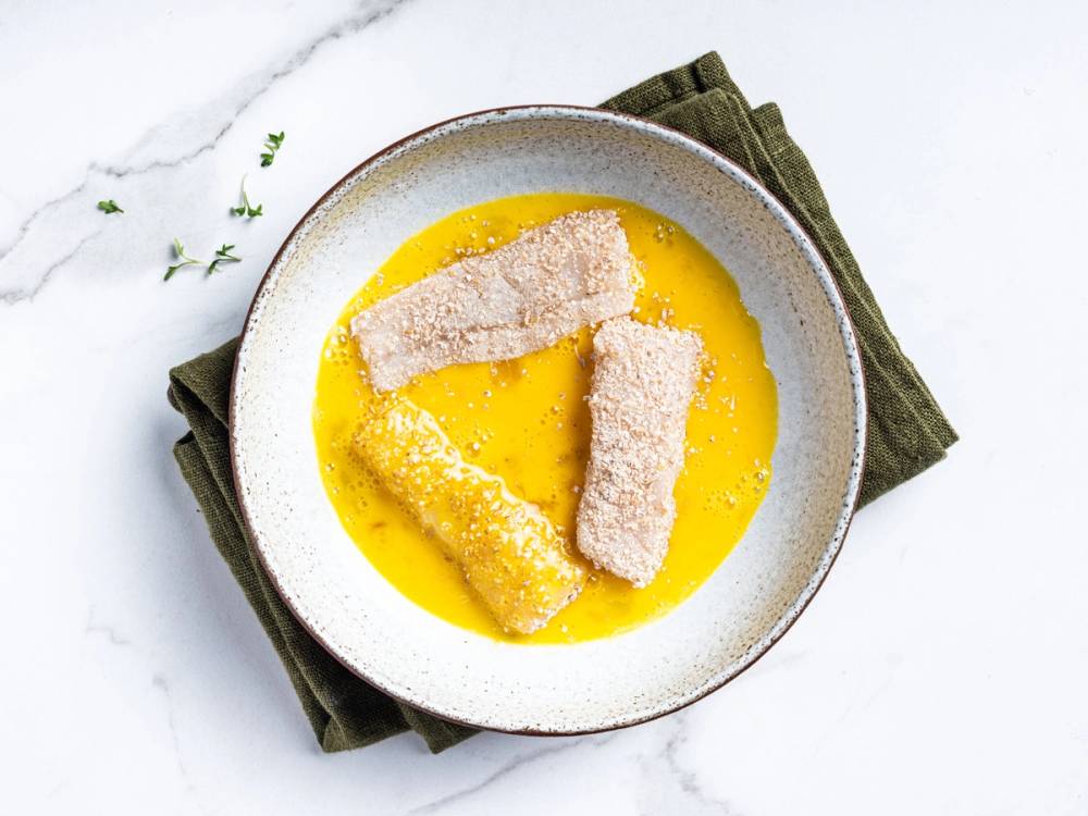 Fiskepinner dyppet i eggeblanding før steking, på en hvit tallerken med en grønn duk under.