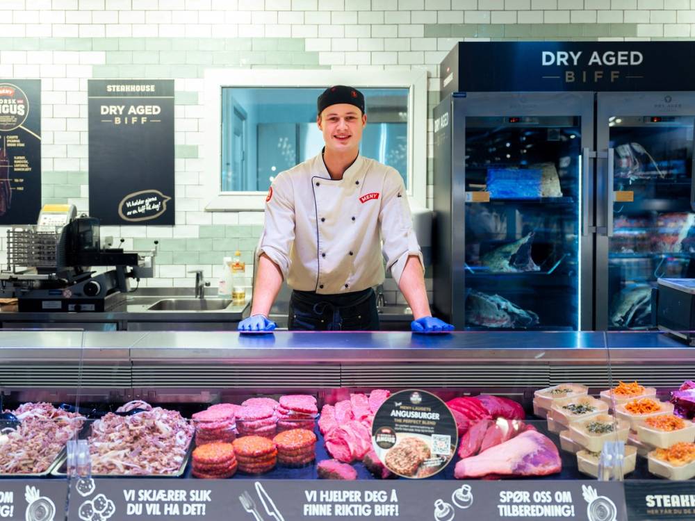 Kjøtt- og pølseprodukter i en kjøttdisk med en ansatt bak disken som har på seg hvit uniform og blå hansker, i en butikk med kjøttavdeling.