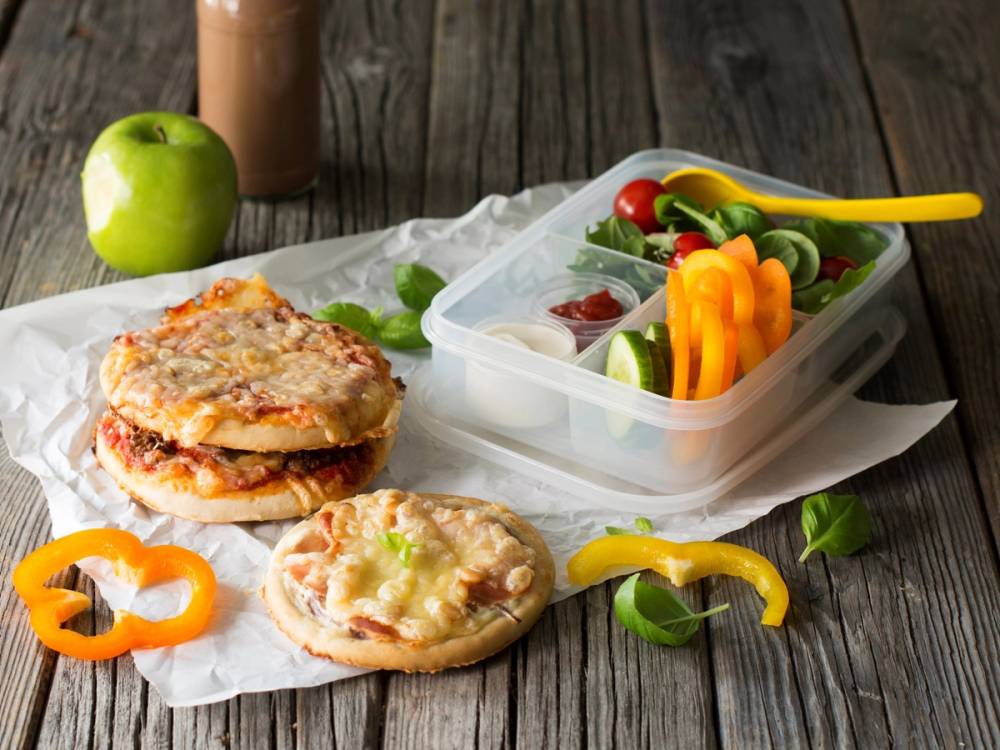 To små pizzaer med ost og tomat, og en boks med grønnsaker og paprika ved siden av, på et treverk.