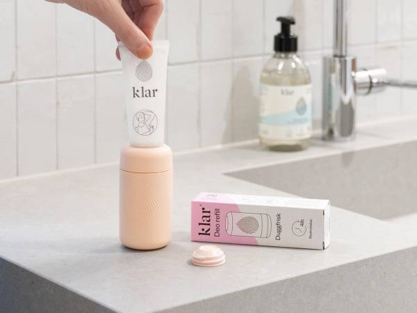 En person holder en hvit deodorant med etiketten 'klar' over en rosa og hvit deodorantboks på kjøkkenbenken.